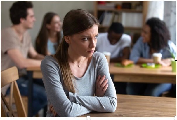 A woman with crossed arms sits at a table looking displeased, while four people converse and smile in the background.