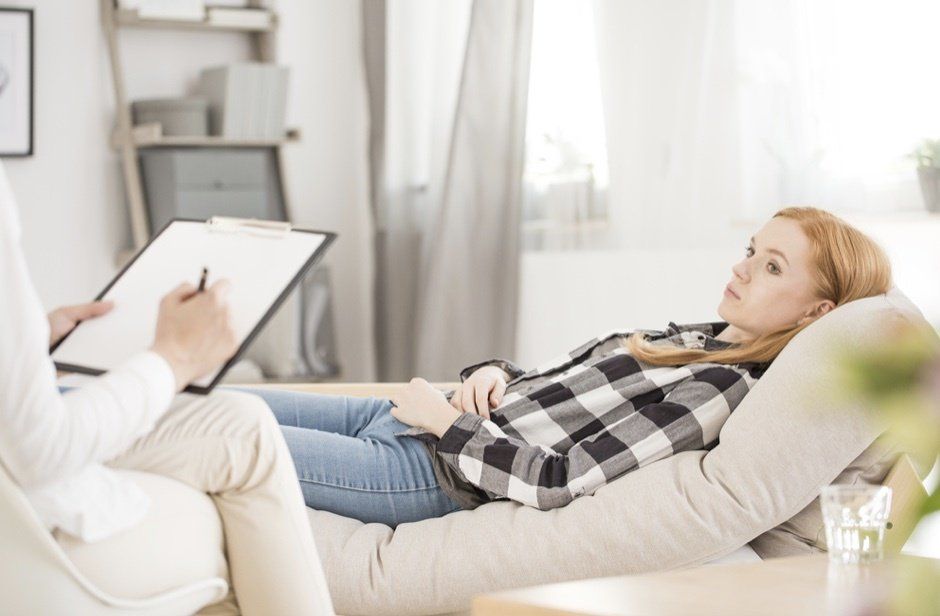 A person is lying on a couch, looking thoughtful, while another person holds a clipboard and takes notes, suggesting a therapy or counseling session in a bright, neutral room.