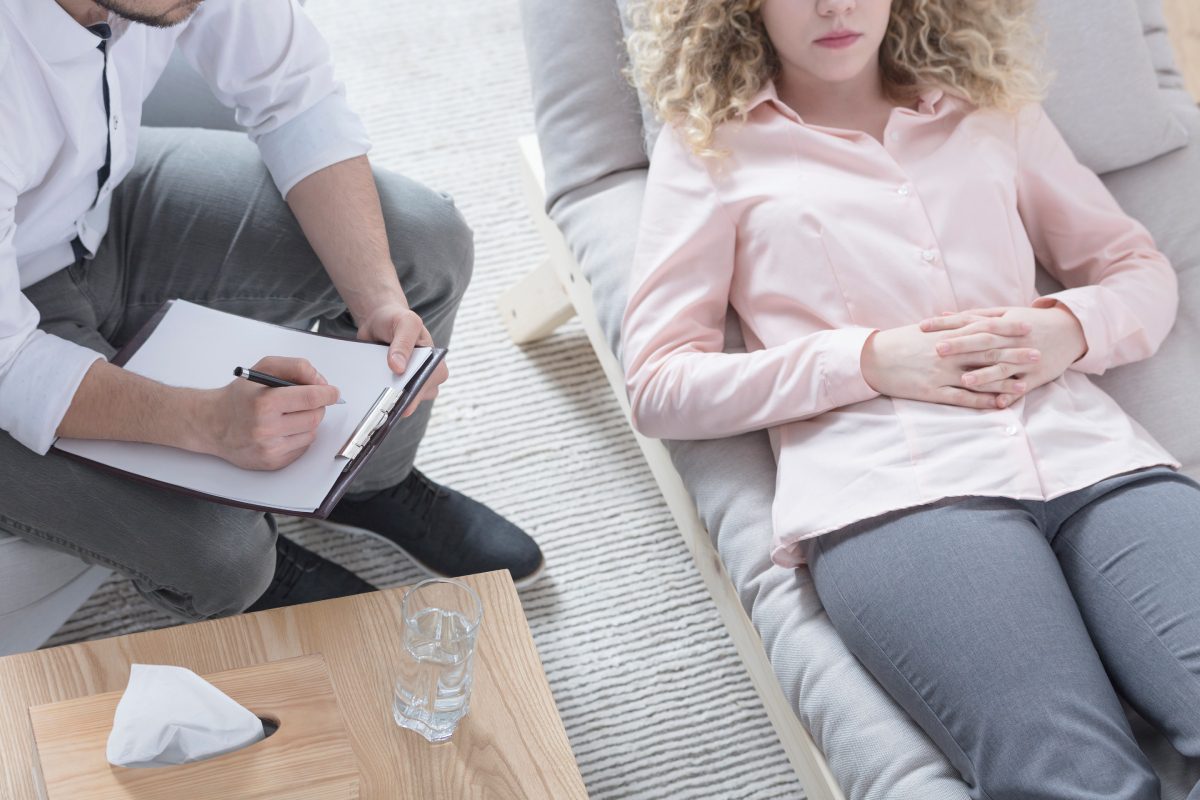 A person lies on a couch during a stress reduction therapy session, while the therapist takes notes on a clipboard. A glass of water and tissues are on a wooden table nearby, creating an atmosphere conducive to relaxation and reflection.