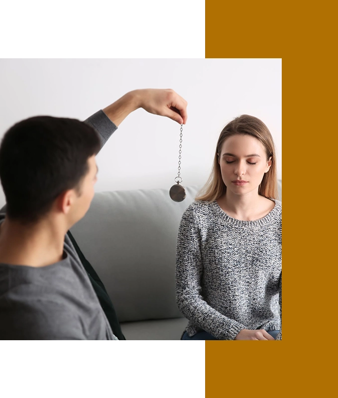 A person is holding a pendulum in front of another who sits on a couch with closed eyes, embracing the calming journey of hypnosis for weight loss.