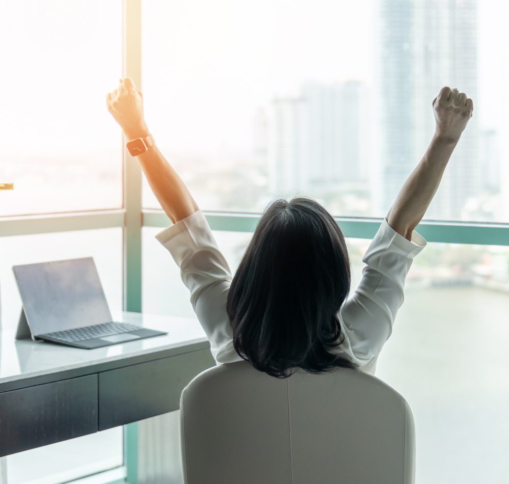 A person sits at a desk with a laptop, raising their arms in triumph. The sense of accomplishment is palpable, enhanced by the emotional wellbeing therapy they've embraced. A stunning cityscape unfolds through a large window, framing their moment of victory.