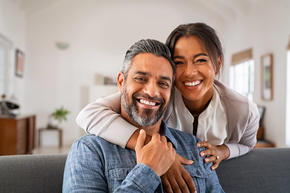 A smiling man with gray hair and beard sits on a couch, embraced by a smiling woman with dark hair, in a well-lit room where the atmosphere exudes warmth and positivity, perhaps benefiting from hypnosis for personal growth.