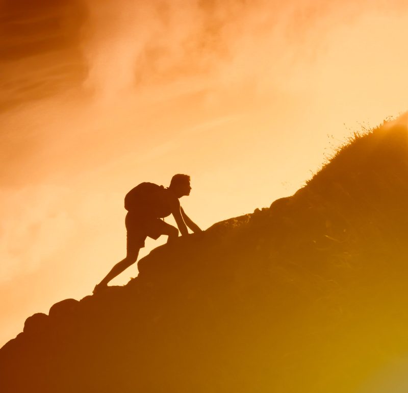 Silhouette of a person climbing a steep hill against a bright orange sky, embodying the journey of hypnosis for personal growth.