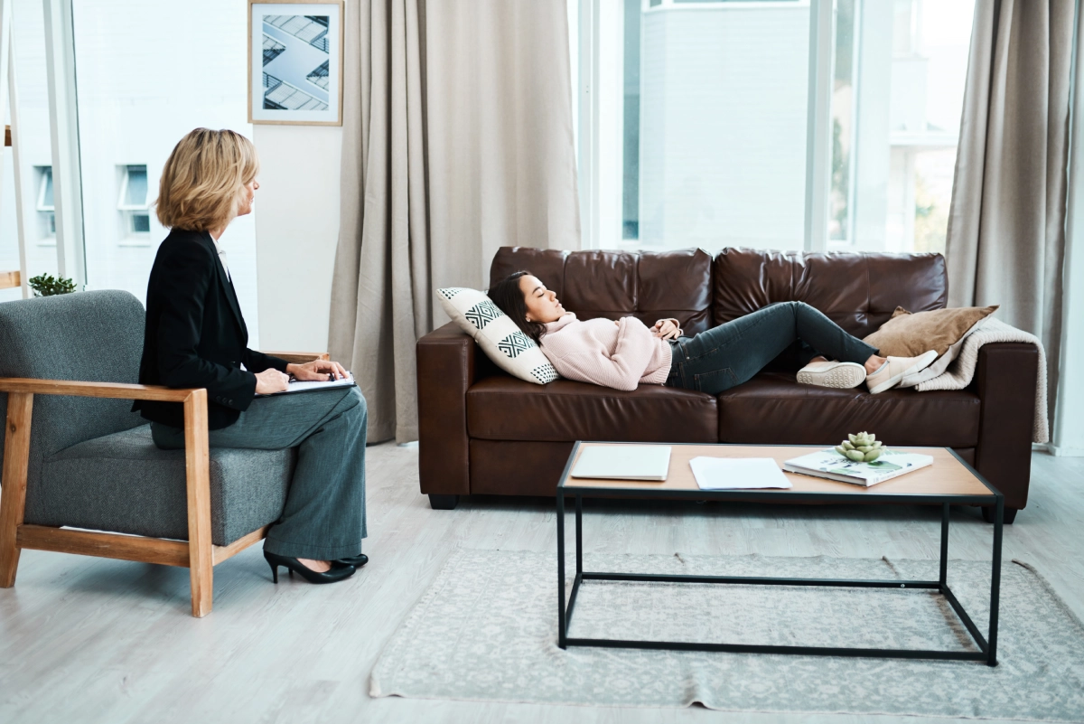 A woman is reclining on a sofa, holding her head as she undergoes hypnosis for anxiety, while another woman sits nearby with a notebook in the modern, well-lit room.