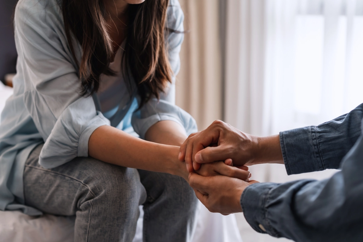 Two people sitting indoors, one holding the other's hand in a comforting gesture, creating an atmosphere akin to a hypnosis session or a comforting EFT practice near me.