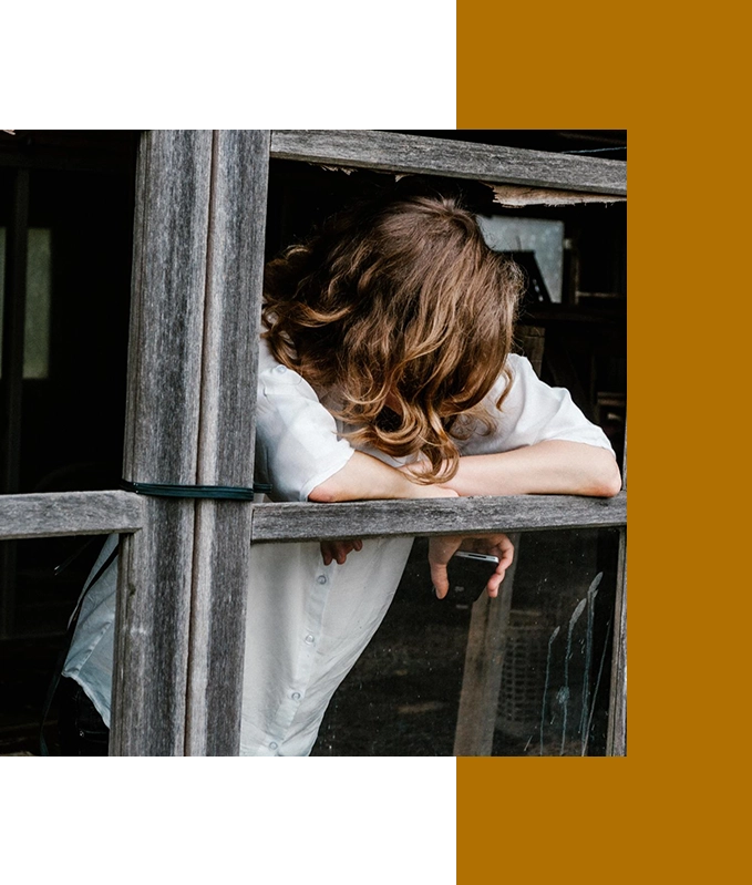 A person with long hair, lost in thought, leans on a windowsill of a weathered wooden structure, their white shirt catching the soft light. The serene moment feels almost like hypnosis, inviting a deep sense of calm and introspection.