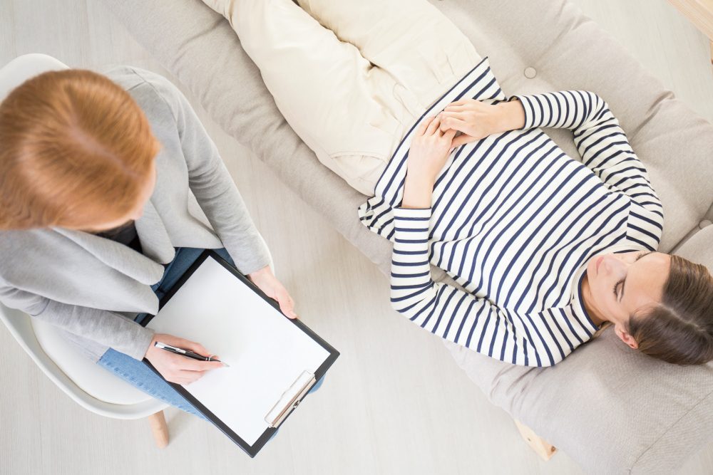 A person lies on a couch while a certified hypnotist sits nearby with a clipboard and pen, suggesting a therapy or counseling session.