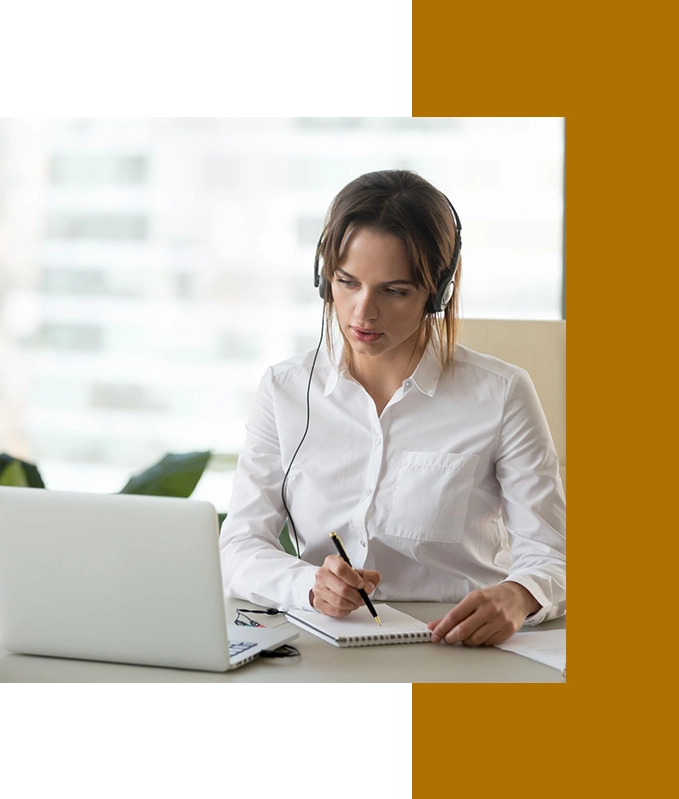 A woman in a white shirt sits at a desk with a laptop, wearing headphones. Engaged in writing, she jots down notes from her EFT Therapy session.