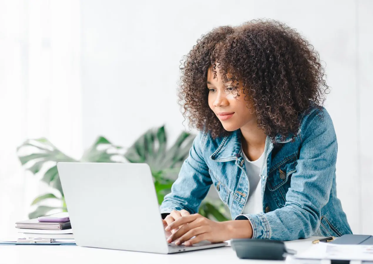 A person with curly hair sits at a desk, engaged with their laptop. They're wearing a denim jacket and are surrounded by lush plants, creating a calming atmosphere that could be perfect for exploring EFT Therapy techniques.