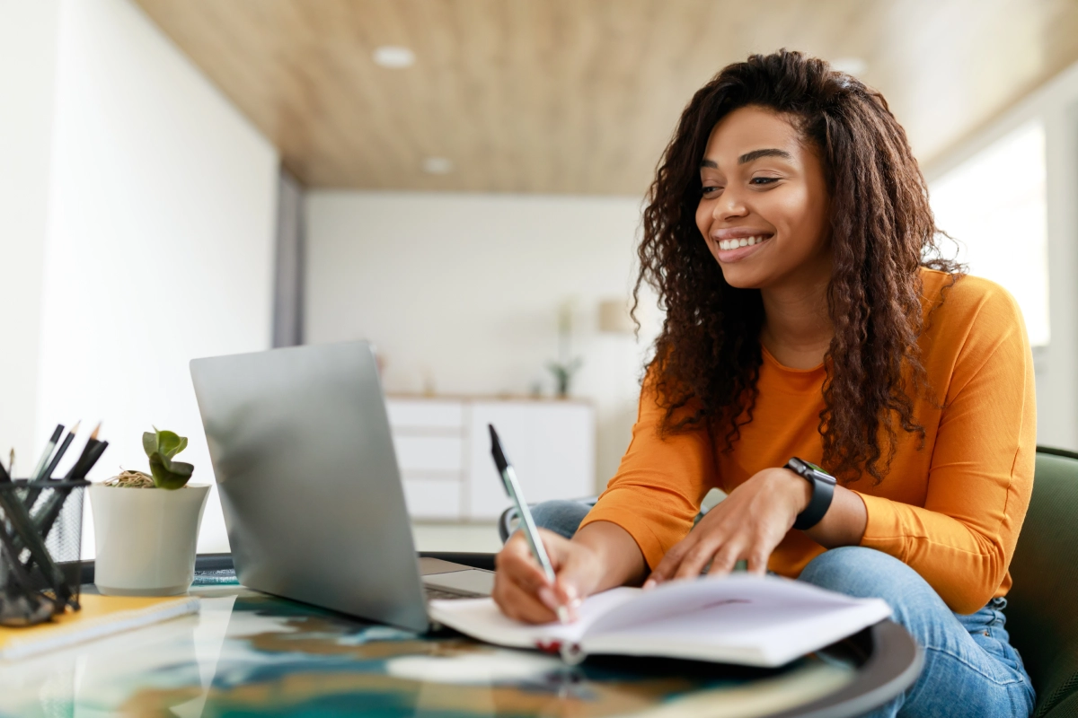 A woman in a yellow sweater is sitting at a table, smiling as she explores EFT Therapy techniques, writing in her notebook and glancing at her laptop screen.