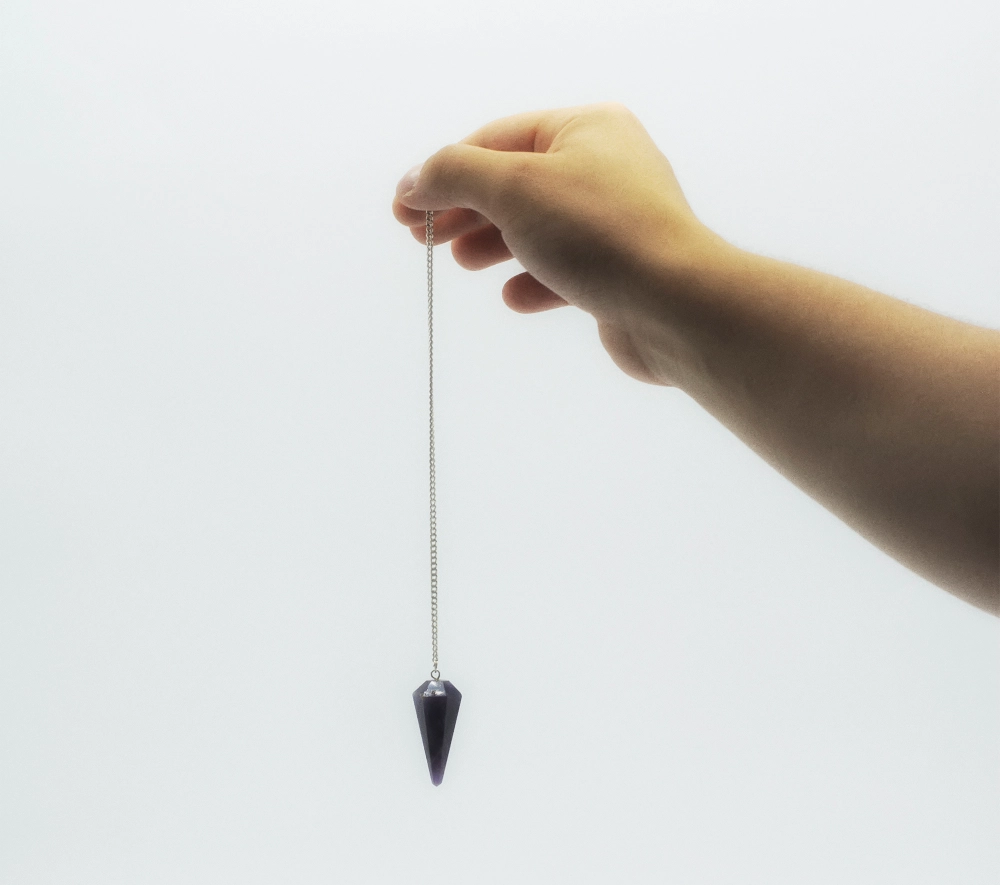 A hand delicately holds a silver chain with a dark, pointed pendant, reminiscent of tools used in hypnosis services in Hartsville, against a plain white background.