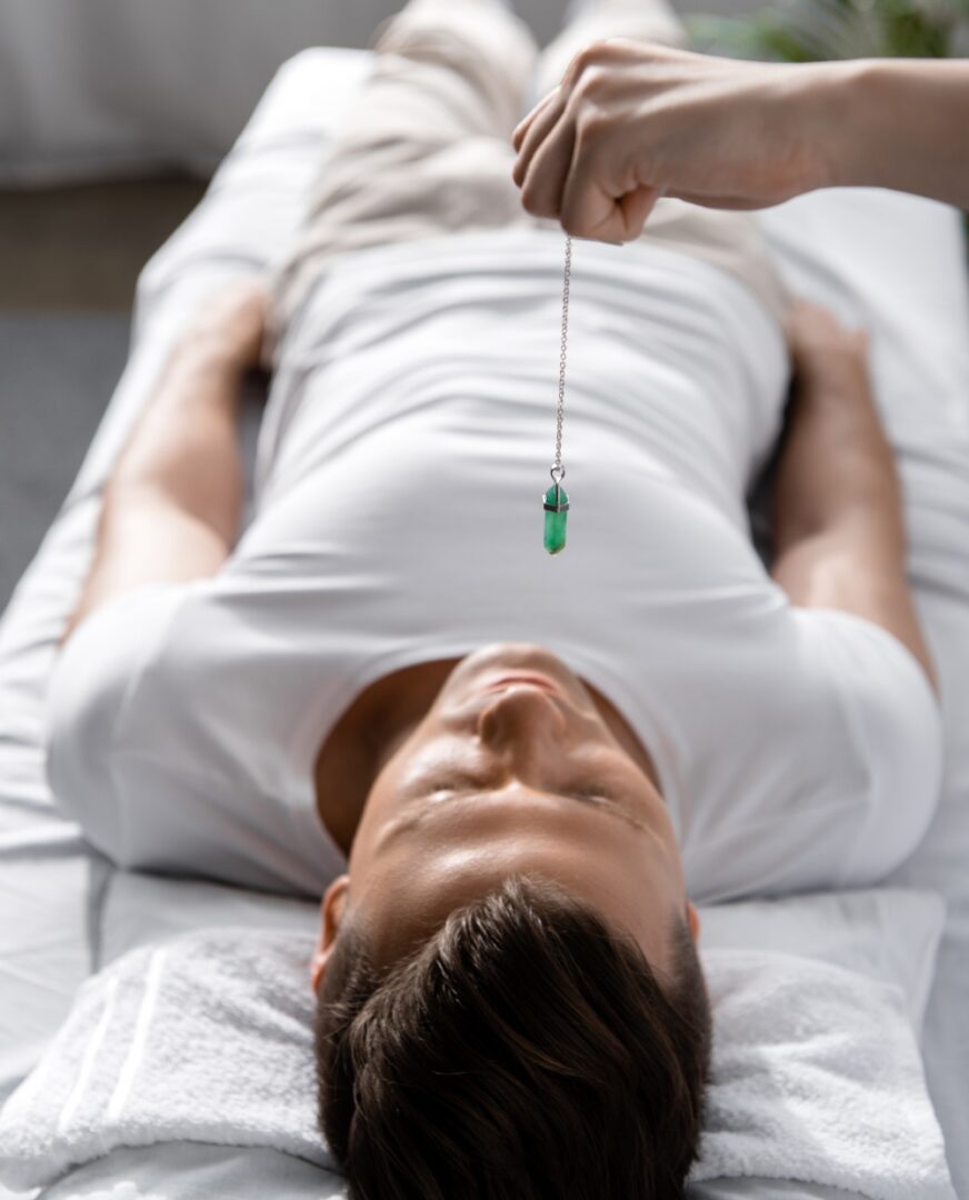 A person lies on a table as another hand holds a green pendant above them, evoking the calming essence of Hypnosis Services in Hartsville.