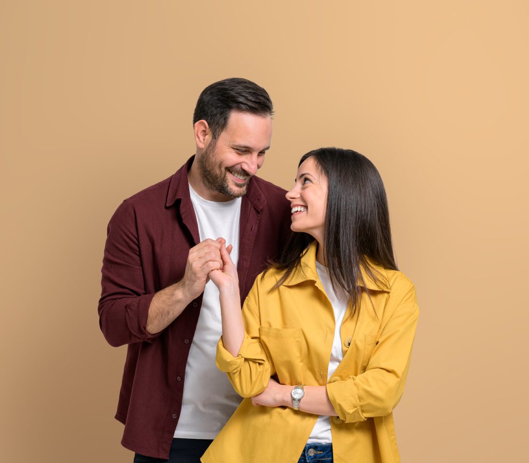 A smiling man and woman stand close together against a plain beige background, radiating confidence. The man wears a maroon shirt, while the woman sports a yellow jacket, perhaps embracing their vibrant journey with Hypnosis Services in Hartsville.