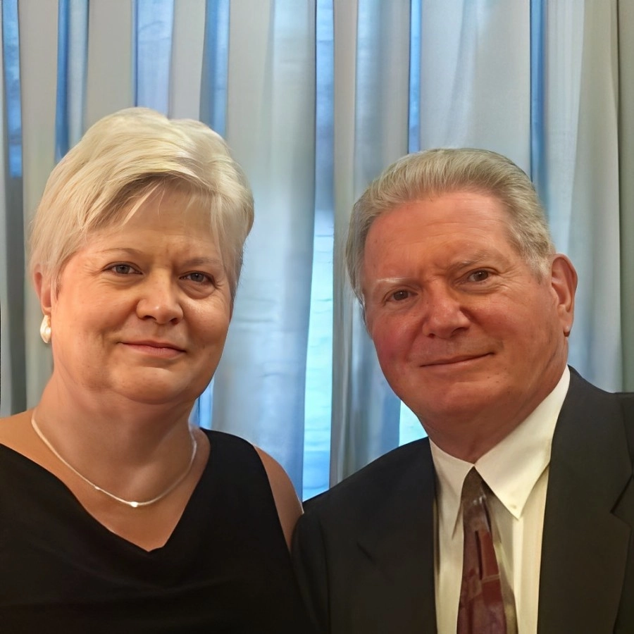 An older couple, dressed in elegant formal attire, stands together in front of a blue curtain. Their serene expressions hint at having discovered profound secrets like hypnosis for weight loss, adding an air of mystery to their poised presence.