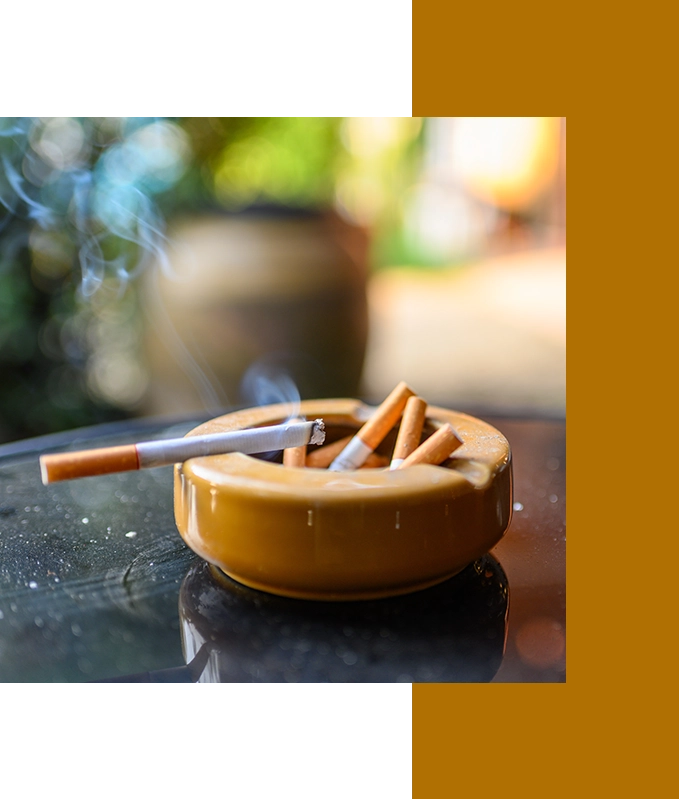 A lit cigarette and several cigarette butts rest in a yellow ashtray on a reflective surface, with smoke swirling like a cloud of thoughts eased by hypnosis for anxiety, set against a blurred background.