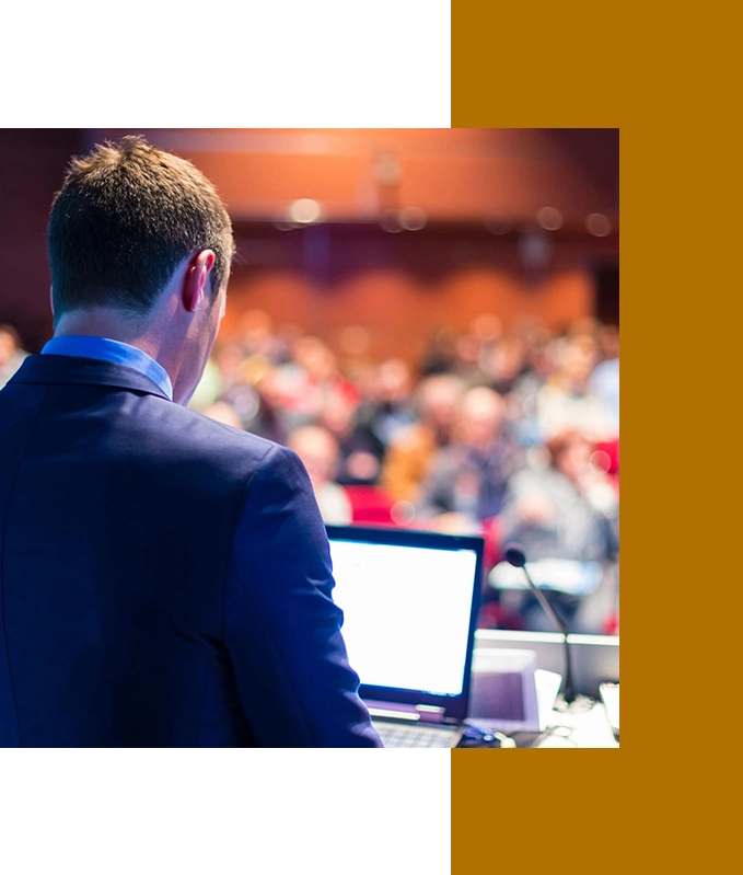 A man in a suit stands at a podium with a laptop, addressing a large, captivated audience in an auditorium, seamlessly weaving in themes of hypnosis for personal growth.