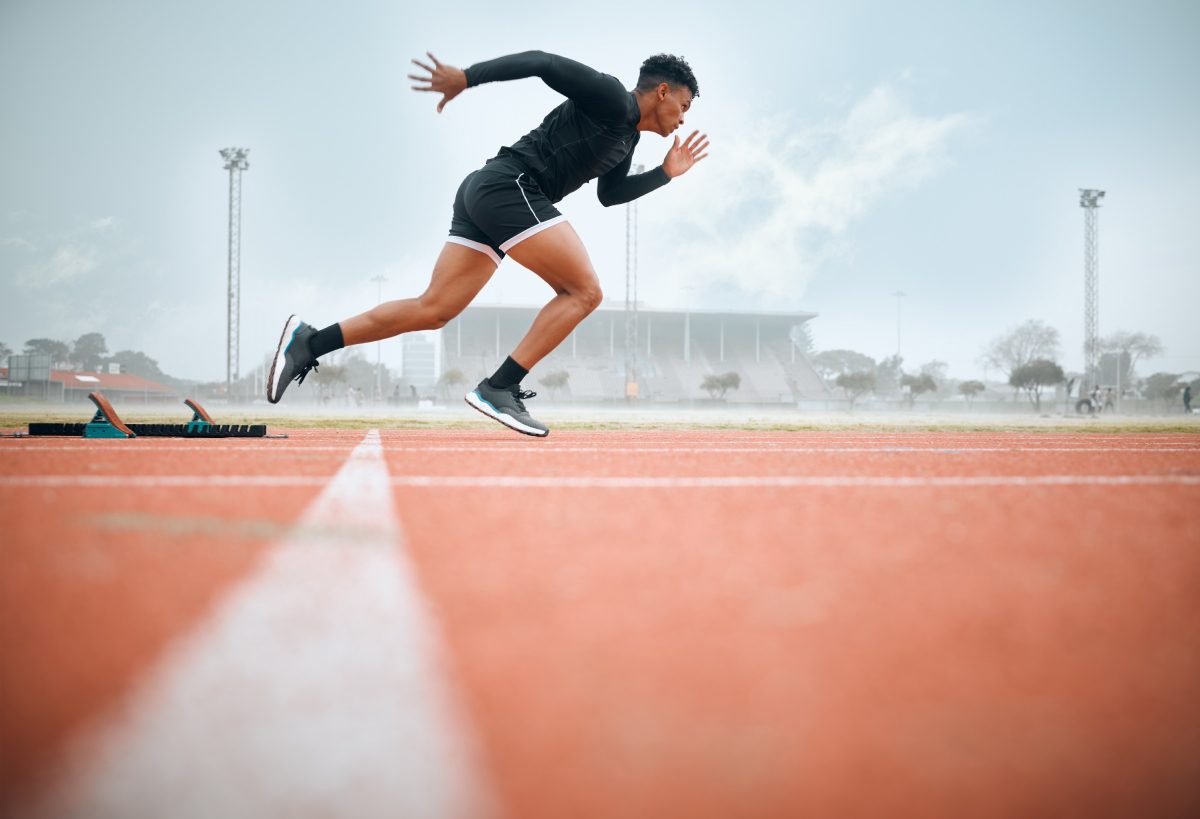 An athlete, fueled by EFT for personal growth, sprints off the starting blocks on a track, with a stadium looming in the background on a cloudy day.