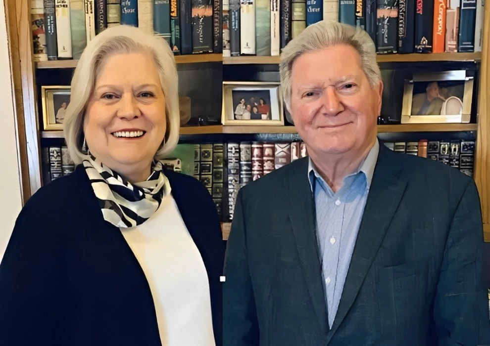 Two people smiling in front of a bookshelf filled with books on wellness and framed photos, reflecting their shared journey through hypnotherapy.