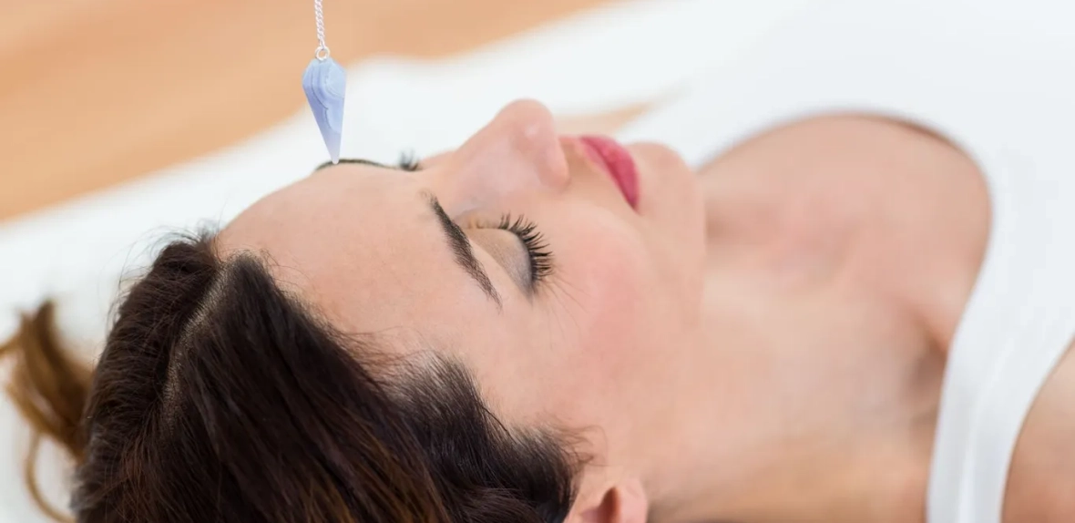 A woman lies down with her eyes closed as a blue pendulum hovers above her forehead, gently guiding her into the realm of hypnosis therapy.