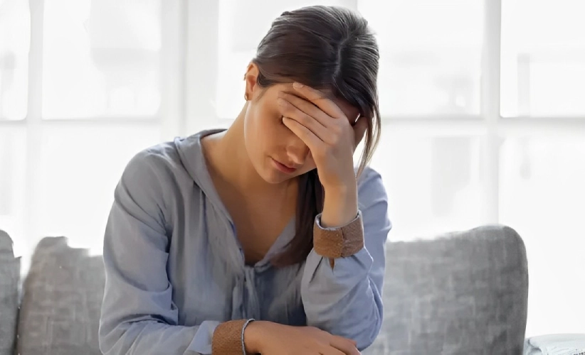 A woman in a light blue blouse sits on a sofa, her elbow on her knee and hand on her forehead, appearing thoughtful or stressed. The serene ambiance and soft natural light suggest a moment of introspection, perhaps contemplating the benefits of hypnosis therapy.