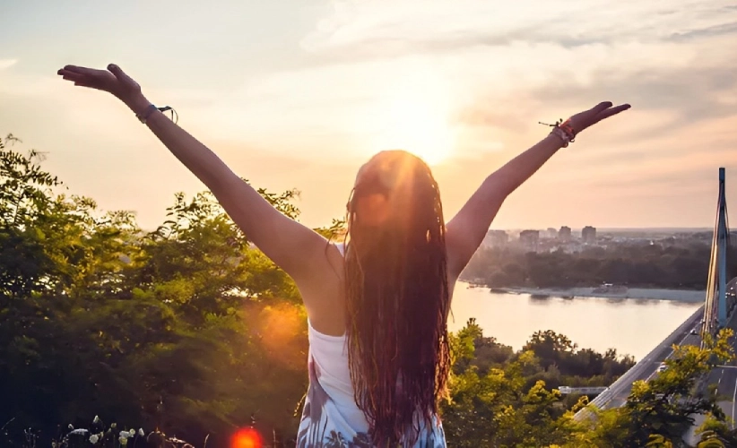 A person with raised arms gazes at the sunset over a river and cityscape, surrounded by trees, feeling as serene as if enveloped in hypnosis therapy.