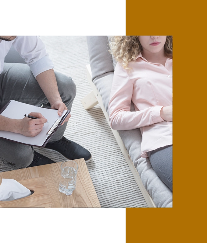 A therapist takes notes while a person lies on a couch, exploring hypnosis for anxiety. A glass of water and a tissue box sit on a wooden table.