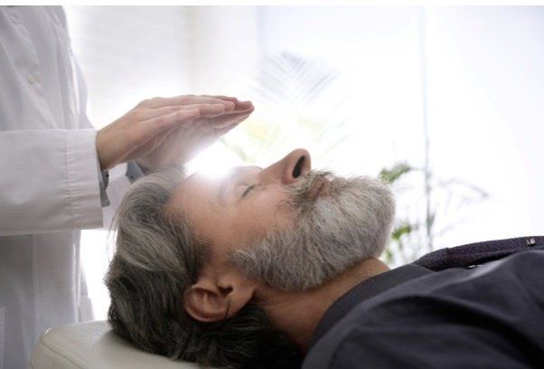 A bearded man lies down with his eyes closed. A person in a white coat holds their hands above his head, suggesting a healing or therapeutic practice.