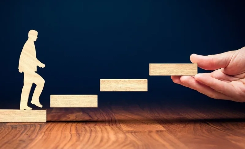 Silhouette of a person walking up wooden blocks, with a hand placing the next block, symbolizing progress or assistance, much like the support and guidance offered through hypnosis therapy.