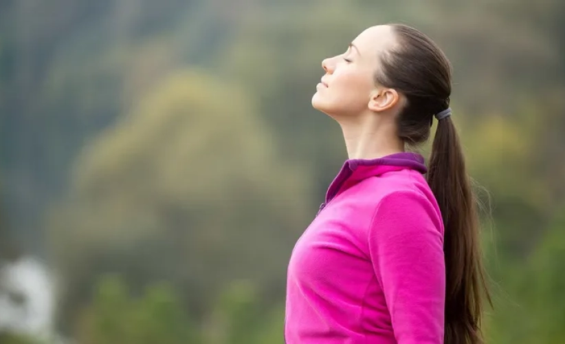 A person in a pink jacket stands outdoors, eyes closed, breathing deeply with a serene expression, as if enveloped in the calming embrace of hypnosis therapy.