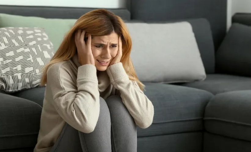 A woman sitting on the floor near a couch, holding her head with both hands, looking distressed. Perhaps she's considering hypnosis therapy to guide her through the turmoil and find peace.