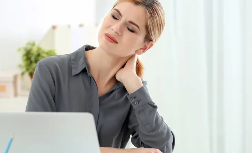 A woman in a gray shirt sits at a desk, eyes closed and hand on her neck, seemingly experiencing discomfort. As the laptop glows beside a small plant, one wonders if she's seeking relief through self-hypnosis therapy.