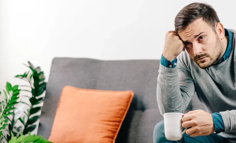 A man sits on a gray couch holding a white mug, resting his head on his fist as if in deep contemplation. Nearby, a plant and an orange cushion create a serene backdrop, reminiscent of a calm space for hypnosis therapy.